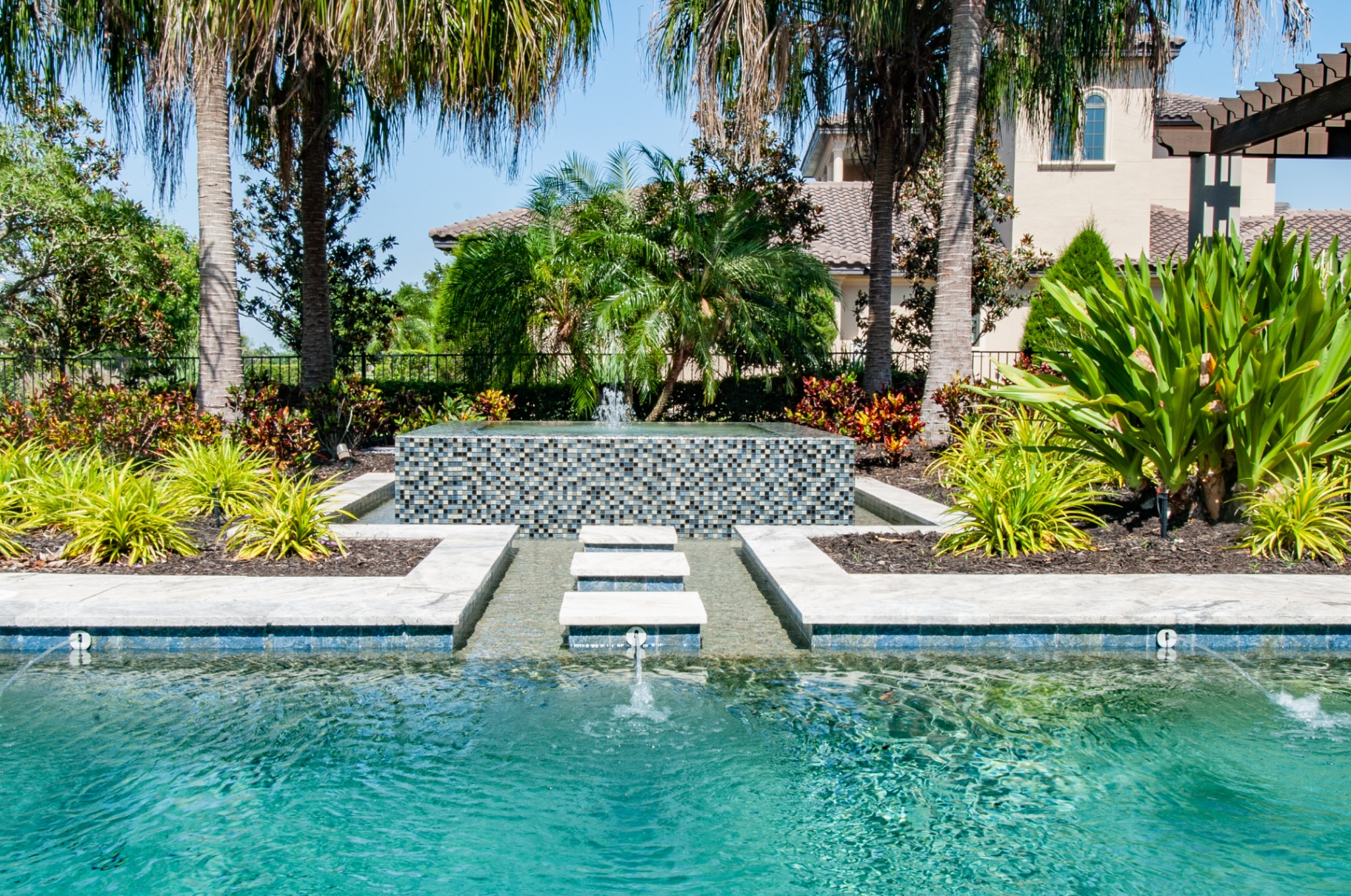Custom mosaic-tile fountain spilling into a turquoise pool framed by tropical plantings
